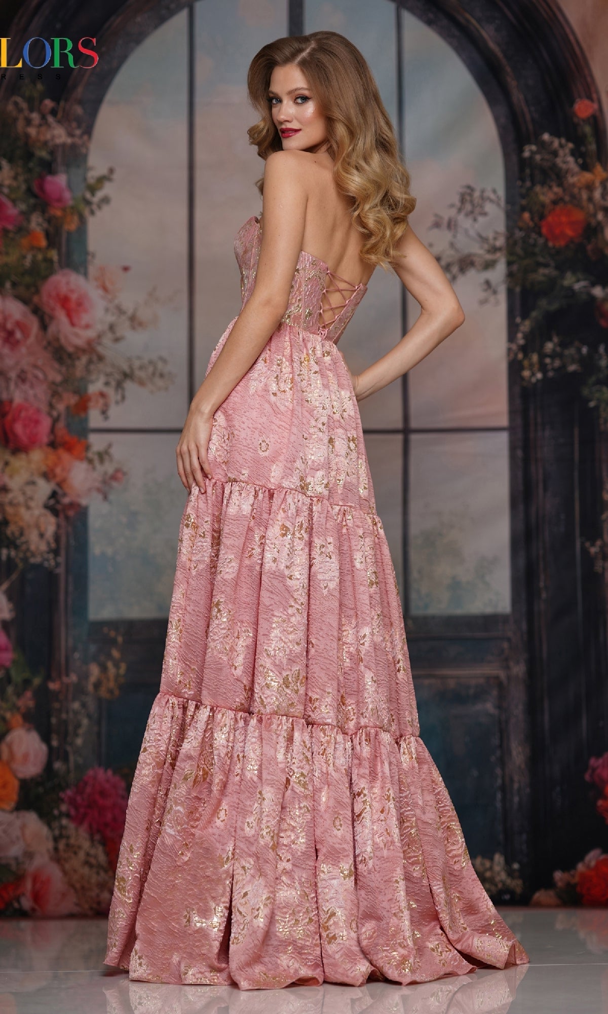 A woman poses with her back slightly turned by a floral backdrop and arched window, wearing the Colors Dress 3794—a strapless long prom dress featuring a corset bodice and tiered flowy skirt.